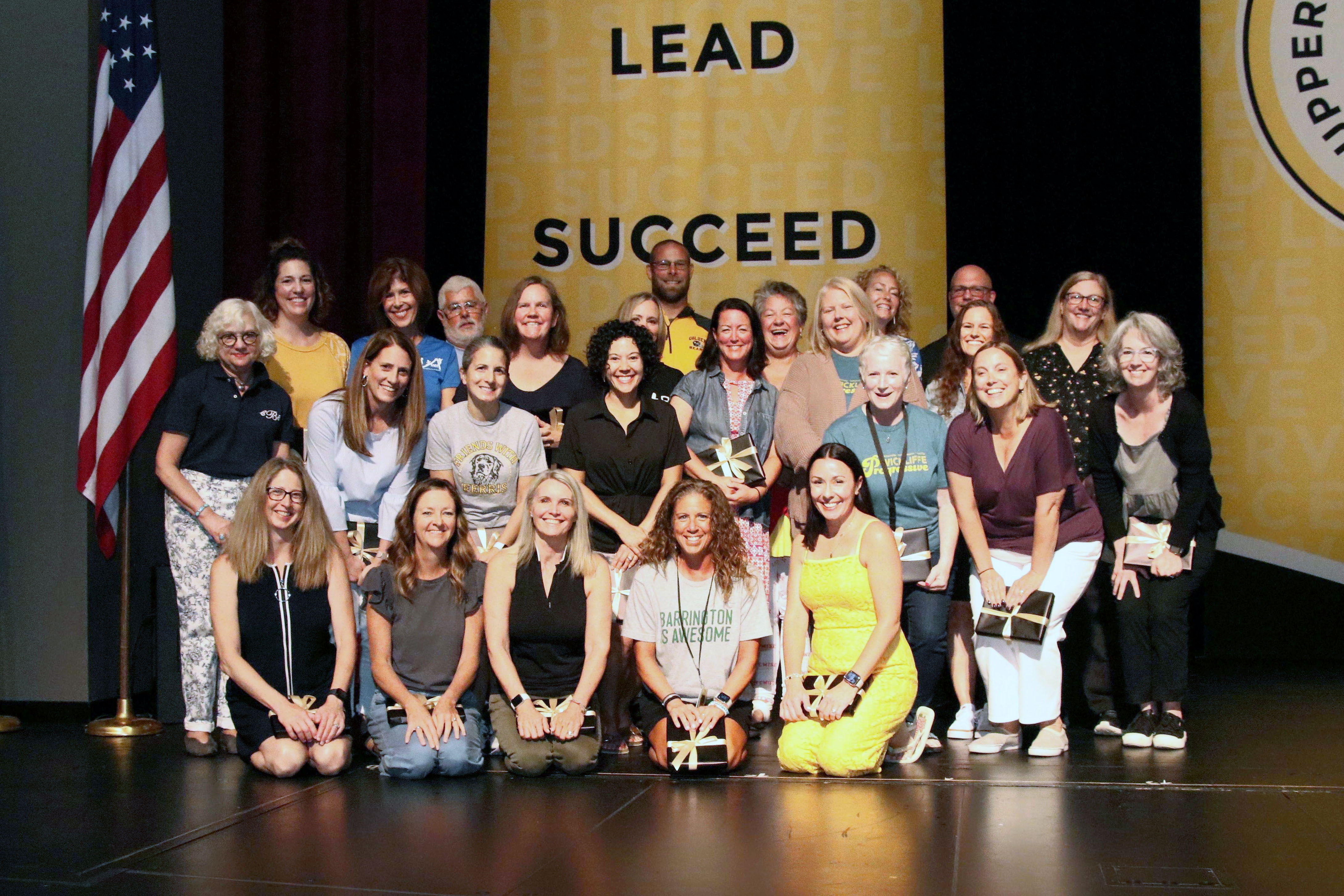 A large group of staff members celebrating 20 years of service with the Upper Arlington Schools posing for a photo on stage with an American flag to the left and gold banners in the background
