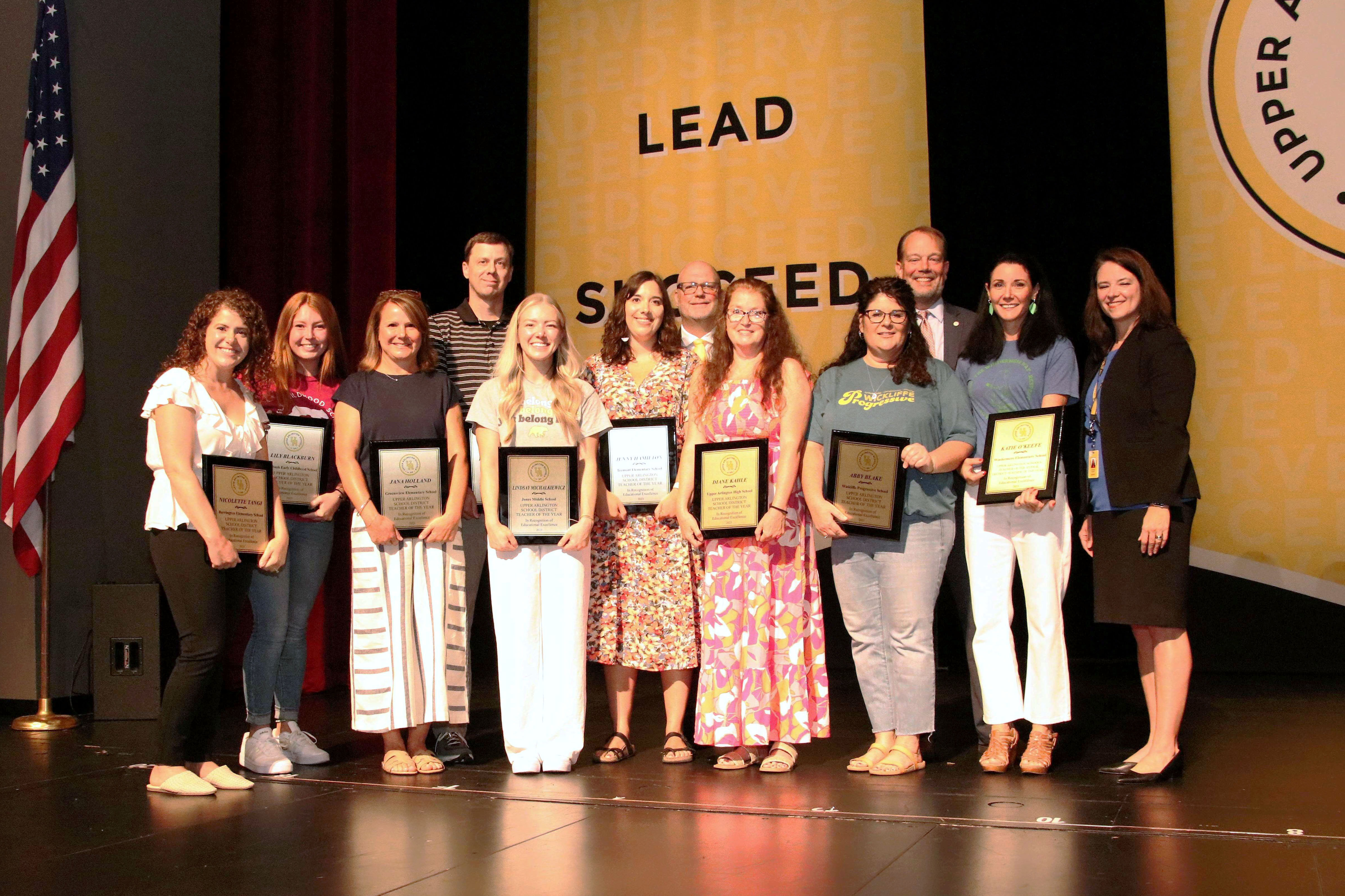 All of the teacher of the year recipients posing on stage with three administrators with gold banners in the background and an American flag off to the left side of the photo