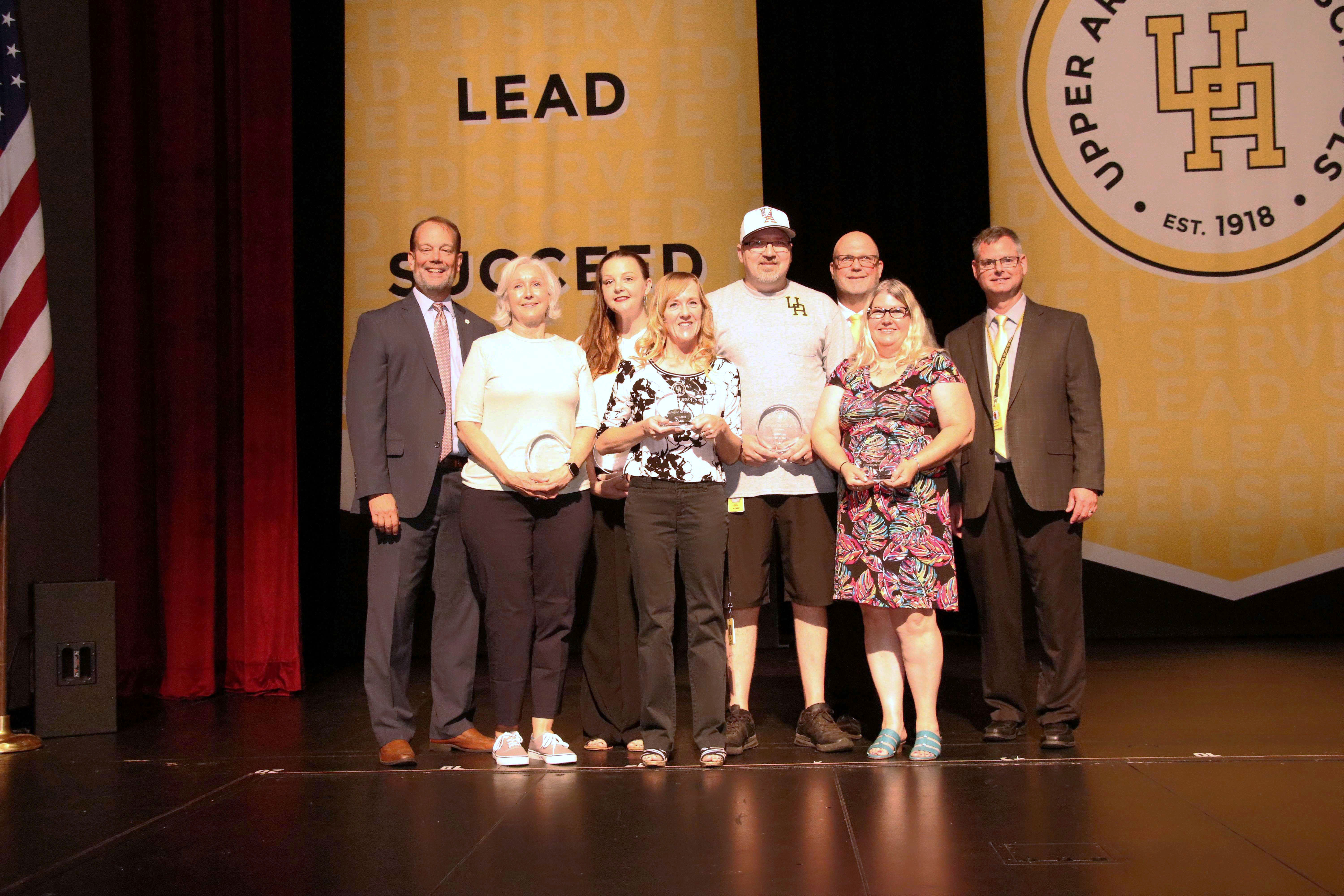 The five recipients of classified and support staff awards posing for a photo on stage with three administrators and gold banners in the background
